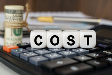 There are cubes on the calculator that say - COST. Nearby out of focus - dollars, notebook and pen