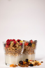 muesli dessert in glass with yogurt and candied or dried fruits with raspberries on top, isolated on white background. raisins, nuts and candied fruits nearby. Granola breakfast