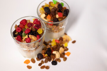 muesli dessert in glass with yogurt and candied or dried fruits with raspberries on top, isolated on white background. raisins, nuts and candied fruits nearby. Granola breakfast