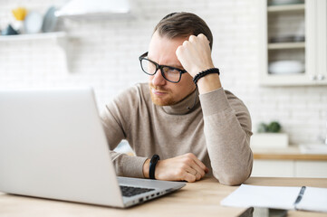 Business man sit at table in kitchen near laptop, resting head on his hand, thinks over strategy,...