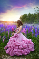 A young girl in a lush pink ball gown with a wild flowers bouquet in her hands against the background of a blooming purple and pink lupines field and a sunset sky.