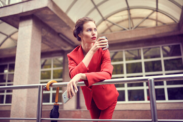 Businesswoman in red costume using mobile phone near office, girl browsing phone, female manager texting smartphone near modern bank building.