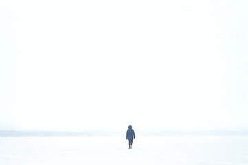 Fototapeta na wymiar a man in a jacket walks through a blizzard on a frozen lake. small person in a large clean space. White sky . place for the test on a white background. matrimony in nature. everything is white.