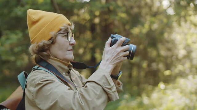 Chest up shot of senior woman with backpack taking picture of nature with professional digital camera while hiking in forest on summer day