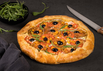 Galette with red and black tomatoes, olives, arugula and ricotta on paper on a dark background 