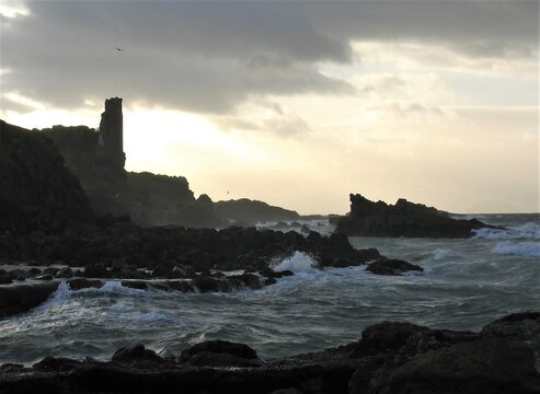Dunure Castle silhouetted against a setting winter sun with waves pounding the coastal rocks during a storm
