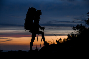 black silhouette of woman with backpack and trekking poles on background of evening sky