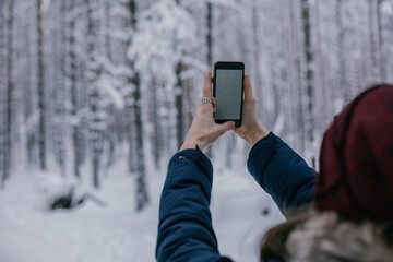 Young beautiful happy girl in red winter knitted hat takes photo in winter forest. Travel and active life concept. Outdoors 