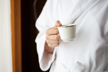 A man in a white terry dressing gown holds a coffee mug in front of a window.