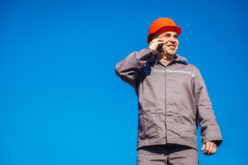 Construction worker in a helmet against the blue sky talking on the phone. Contractor control according to plan. Builder engineer helmet works at construction site. Copy space.