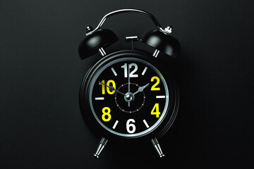 A stylish black alarm clock with yellow and white numbers stands on a dark background. The concept of the transience of time. Blank for the designer