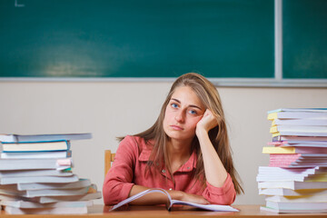 Teacher surrounded by books sitting in school classroom.Emotional teacher at the table. Copy space. Funny photo. Beautiful young girl.Many study tasks