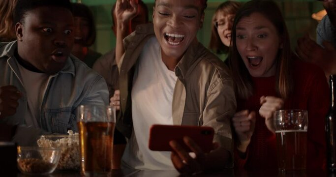 Cheerful afro american woman holding phone horizontally and rejoicing while sitting with friends in pub and looking at screen. Group of diverse excited people watching sport at smartphone