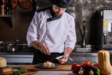 Italian cook adding spices to dish on plate, young professional chef in uniform decorating...