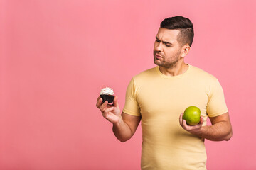 Joyful young Caucasianman in casual made wrong choice deciding between cake and fresh green apple....