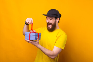 Amazed bearded hipster man opening a gift box over yellow background.