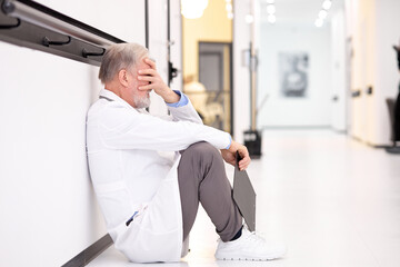 Tired exhausted male senior doctor sits on hospital floor alone. Depressed sad caucasian doctor feels fatigue burnout stress, lack of sleep, napping at work