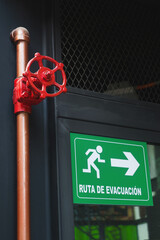 Una válvula roja y un tubo largo al lado de una reja oscura, un espejo y un aviso verde de ruta de...