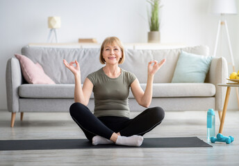 Balanced senior woman sitting in lotus pose and making gyan mudra with her hands, meditating on sports mat at home