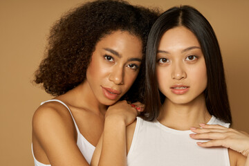 Beauty portrait of two mixed race young women with perfect skin looking at camera while posing together, standing isolated over light brown background