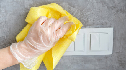 female hand clean light switch with cloth on gray concrete wall. Woman in glove disinfecting...