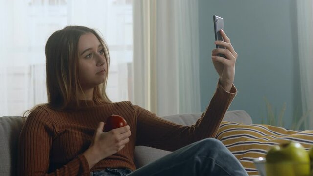 Beautiful woman in blue jeans and brown sweater sits on beige sofa on window background, holding red apple and taking selfie. Average static plan.