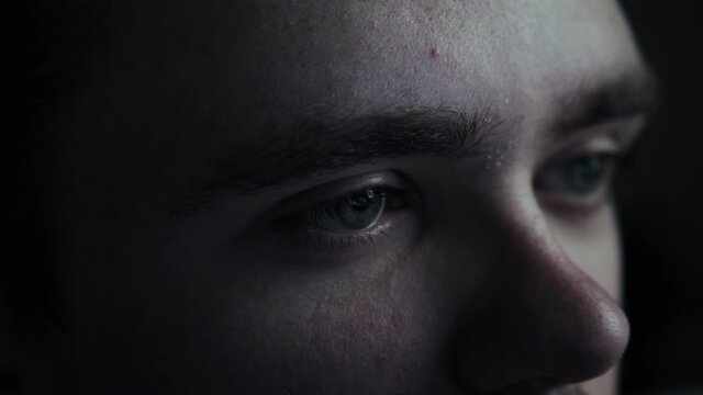 A close-up of a young guy sitting in a dark room watching TV with fright and fear. Focus on one side of the face. Weekend and relaxation concept