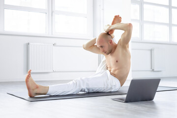 Naklejka premium shirtless yogi man showing yoga exercises at laptop camera, teaching online via Net, webcam. man sitting on floor , flexible and sportive