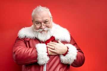 santa man 60 years old expressing attitude, holding hands on chest and looking at camera, wearing red winter coat. isolated red background