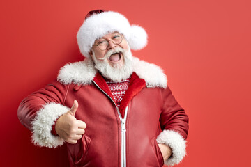 senior santa in red coat and hat shows thumbs up isolated over red studio background, handsome happy man ready to please people, happy with the result