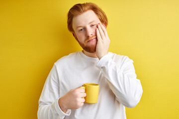 Dissatisfied male student need some sleep, stand with cup of coffee isolated on yellow studio background, portrait. Young guy in casual white shirt has bad mood