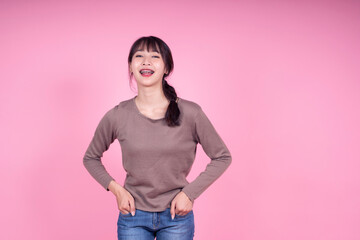 Beautiful young attractive Asian girl woman happy smiling laughing with retainer braces good dental hygiene, casual clothing hands in pocket studio copy space pink isolated background advertisement
