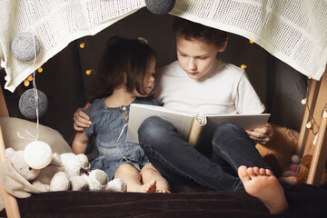 Siblings sit in a hut of chairs and blankets. Brother and sister reading book with flashlight at...