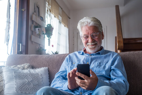 Old Man Smiling Sitting On The Sofa In The Living Room Holding Phone, Enjoying Using Smartphone Feeling Satisfied Sending Messages, Calling Friends, Surfing Web Online Concept