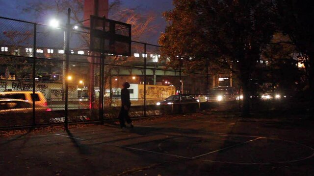 Man in Beanie Plays Basketball Outdoors, NYC Subway Train Moves at Night on Bridge in Background - 02