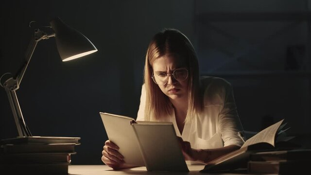 Night learning. Hard task. Procrastination problem. Confused tired troubled female student in eyeglasses doing homework disappointed stopped reading closing textbook late at dark home workplace.