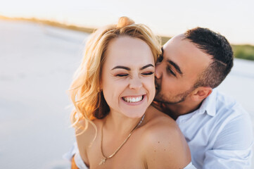 Fototapeta premium A bearded man and a blond woman hugging against the background of white sand. Portrait of emotional people at sunset. Love in the desert newlyweds. The love story of merry lovers young.Kiss and smile.