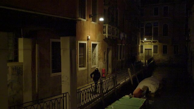 Pedestrian on sidewalk next to narrow water canal in Venice at night