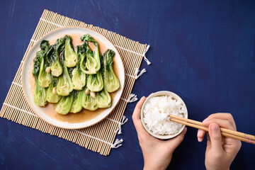 Stir fried bok choy with soy sauce on plate eating with cooked rice by using chopsticks on blue background, Asian vegan food