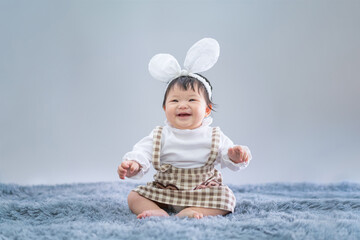 Baby in Easter bunny hairband. Asian happy baby smiling and sitting on carpet on gray background. Cute 6 months baby start sitting with copy space as Easter concept, baby or kid department in hospital