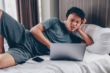 Young Asian man is sad and angry when playing a game in laptop and phone at bed