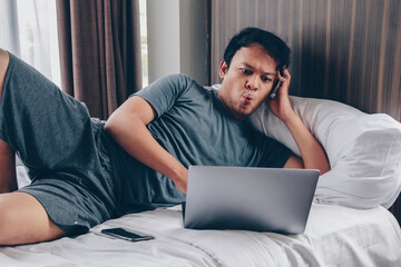 Surprised and shocked Asian man is working with his laptop on his cozy bed. Concept of successful freelancer lifestyle.