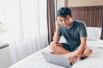 Tired stressed young Asian man feeling sleepy and tired while using laptop on the bed in bedroom. Hard work concept.