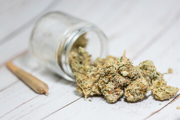Trimmed cannabis buds in glass jar with rolled marijuana joint on white wood