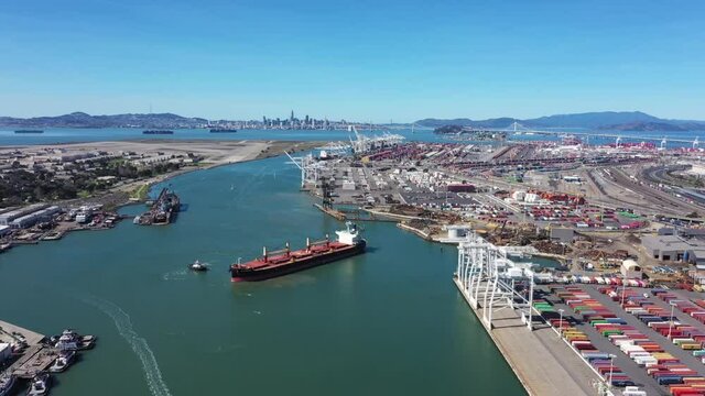 Container Ship being turned around by tug boats on the Oakland inner bay. Container cranes nearby and San Francisco in the background on a clear day. Shot from above and moving towards the ship.