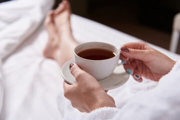 a cup of tea in a soft bed. a young woman in a white coat drinks morning tea