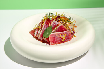 Tuna tataki in sesame with salad. Fish sashimi restaurant appetizer on white table with green wall. Day sunlight with hard shadow of tropical palm leaves. Summer or spring food concept.