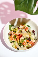 Poke bowl with shrimp and vegetables. Restaurant seafood appetizer on white table with pink wall. Day sunlight with hard shadow of monstera palm leaves. Summer or spring restaurant food concept.