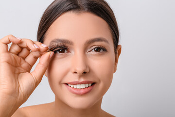 Beautiful young woman applying fake eyelashes against light background