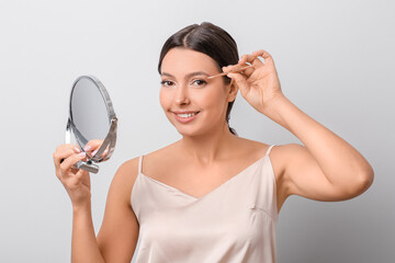 Beautiful young woman applying fake eyelashes against light background